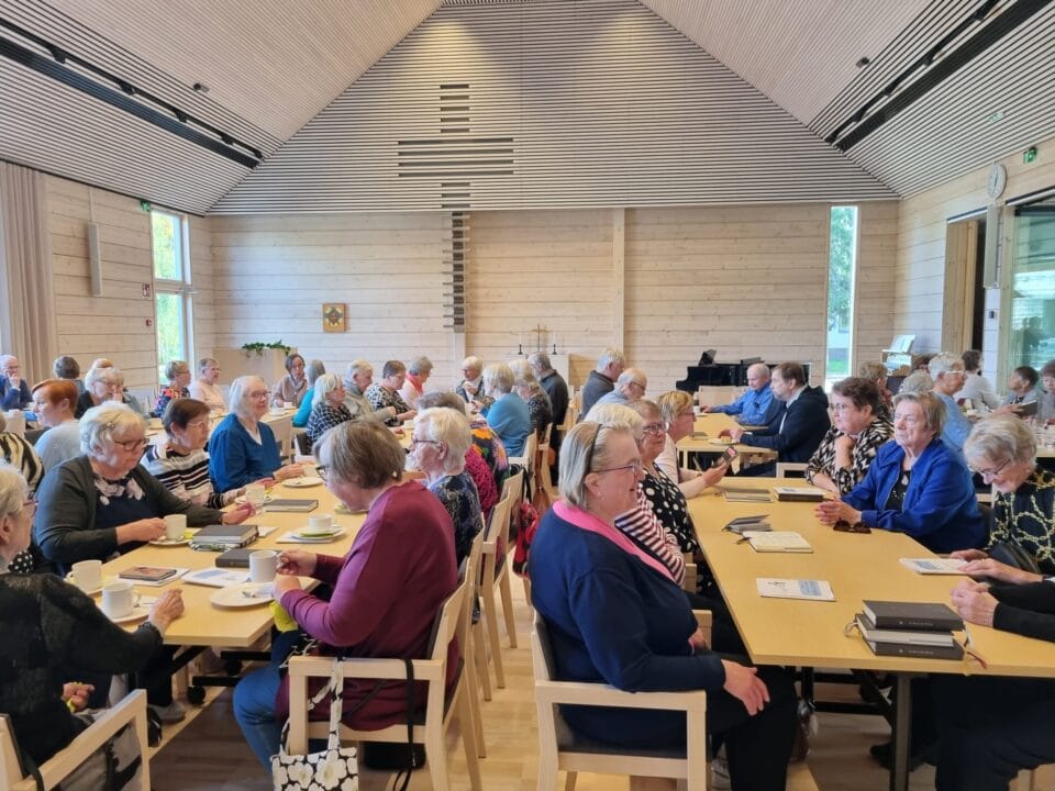 Kuvassa seurakuntakodin usean pöydän ympärillä istuu kahvittelemassa runsaslukuinen osallistujajoukko